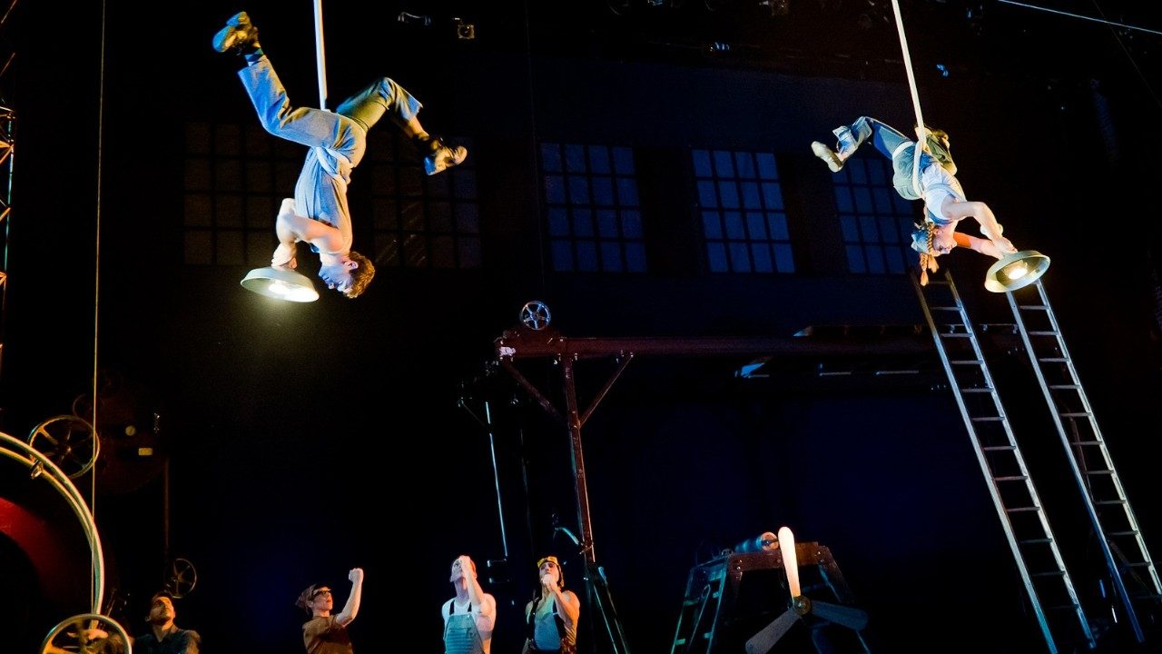  Two members of Cirque Mechanics hang upside down from lights on stage while other members look on.
