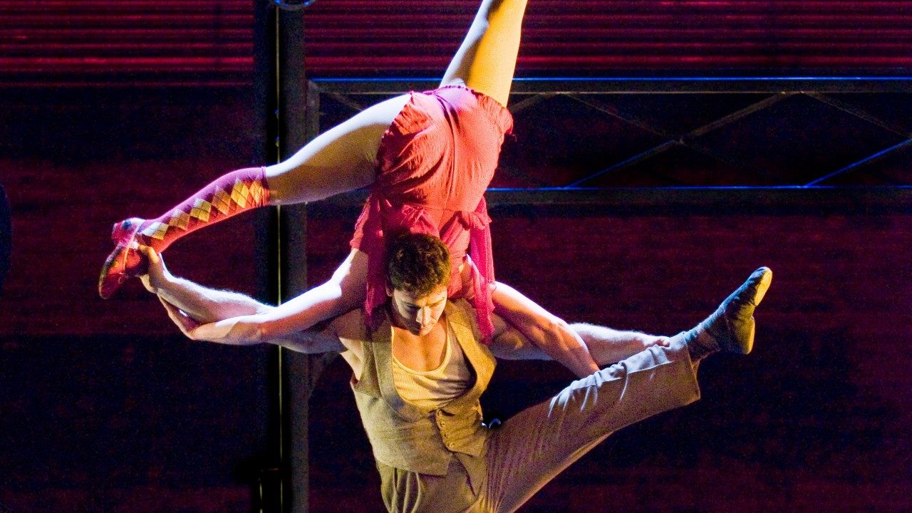  Two members of Cirque Mechanics: a man stands with one leg raised. Perched upside down on his shoulders is a woman.