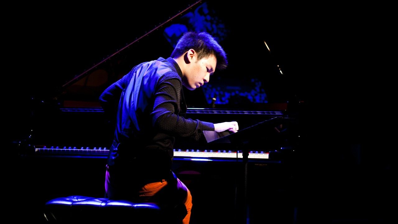  Conrad Tao, an Asian man with dark hair, plays the piano in a dark shirt and reddish orange pants. He is lit from above by a blue light.