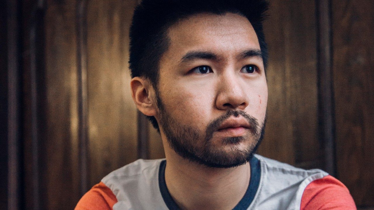 Conrad Tao, an Asian man with dark hair and a closely trimmed beard, sits in front of a warm brown wooden paneled wall, wearing a light grey T-shirt with a navy blue trim at the neck and orange sleeves