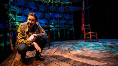  Actor DeLanna Studi crouches on a hexagonal wooden deck in a brown jacket and dark pants during her solo play, "And So We Walked," about the Trail of Tears