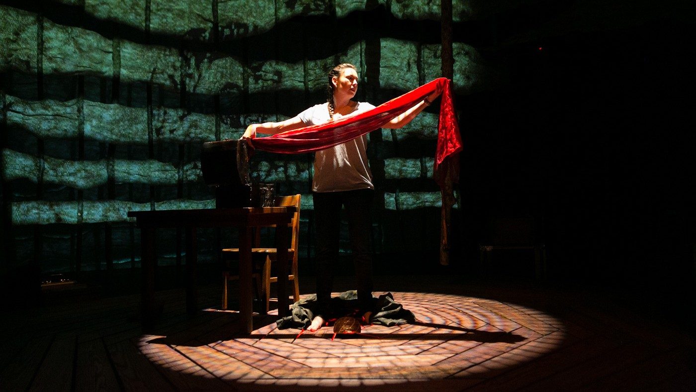  Actor DeLanna Studi stands in the middle of wooden decking and holds a long red scarf between her outstretched hands 