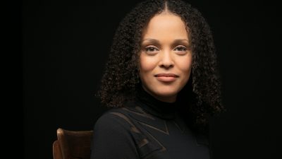 Jesmyn Ward, a light skinned black woman with shoulder length natural hair parted down the middle, smiles towards the camera in a black sweater against a black background.