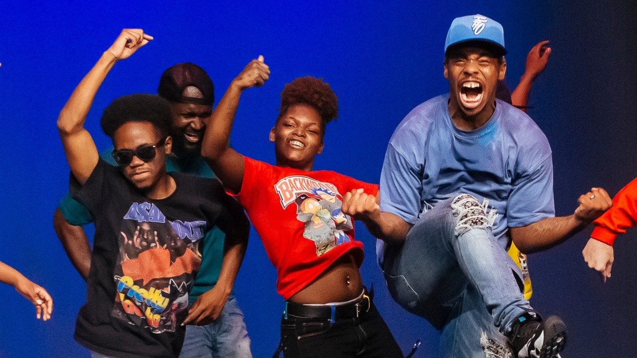  Some of the cast of "Memphis Jookin': The Show" dance on stage. In the foreground, Lil Buck, a Black man in a blue T-shirt, blue hat, jeans, and sneakers, jumps up into the air and pulls his upturned fists in towards his sides in celebration. He's screaming with joy and excitement. Behind him, other dancers, Black men and woman in jeans and T-shirts, hit poses with their hands up in the air above them.