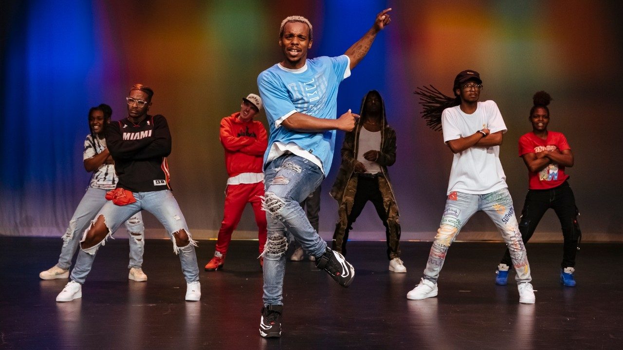  Cast members of "Memphis Jookin': The Show" dance on stage. The background is lit with multicolored lights. In the foreground is Lil Buck, a Black man with short, natural bleached hair, a light blue T-shirt, ripped jeans, and sneakers, dances. He is on one foot with his other bent at the knee, his arms swinging as he dances. Behind him, Black men and women in T-shirts and jeans dance to choreography, all hitting the same pose with their stance wide and their arms crossed in front other their bodies.
