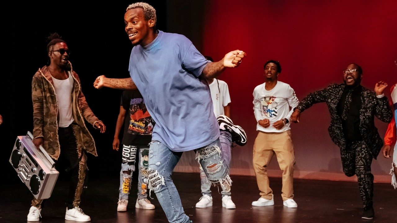  Cast members of "Memphis Jookin': The Show" dance on stage. The background is lit with red lights. In the foreground is Lil Buck, a Black man with short, natural bleached hair, a light blue T-shirt, ripped jeans, and sneakers. He dances; he is on one foot with his other bent at the knee behind him, his arms bent to the sides. Behind him, Black men and women in T-shirts and jeans dance and cheer.