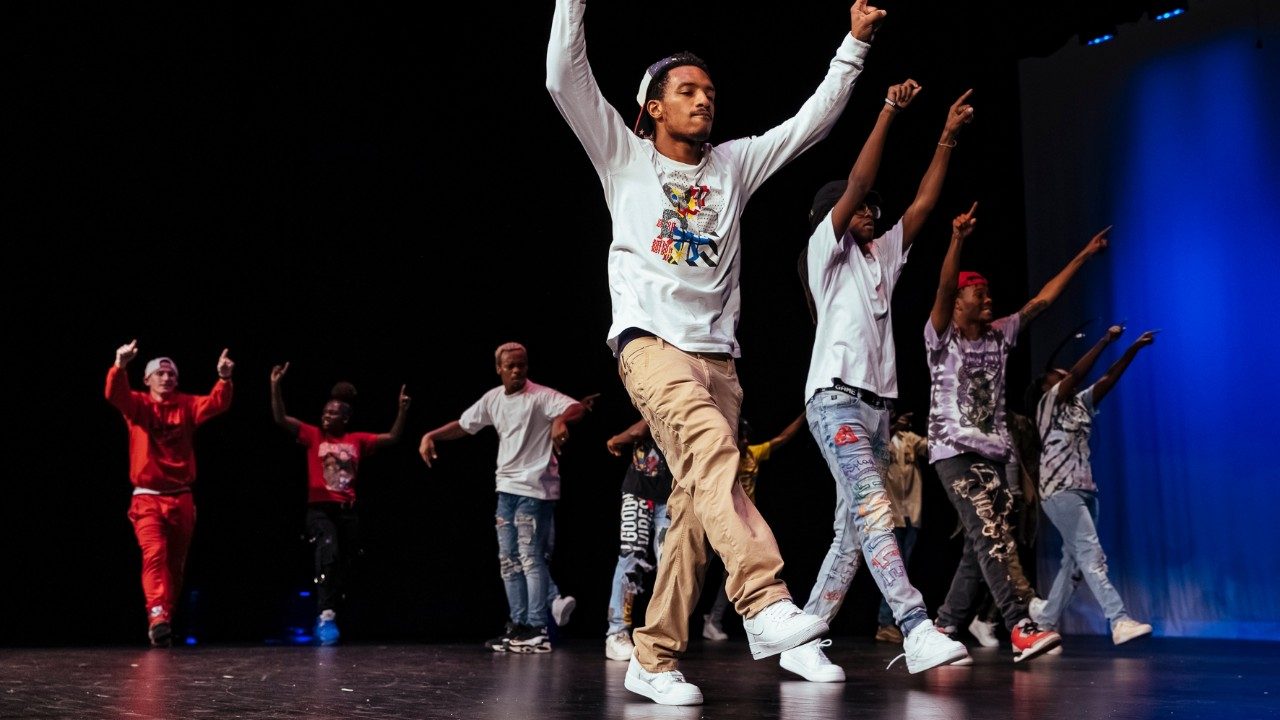  Some of the cast of "Memphis Jookin': The Show" dance on stage. The background is lit with blue lights. Four Black men in T-shirts, pants, and sneakers walk in a line towards stage left, each stepping forward on their right legs and with both arms raised above their heads. Behind them, other members dance and cheer.