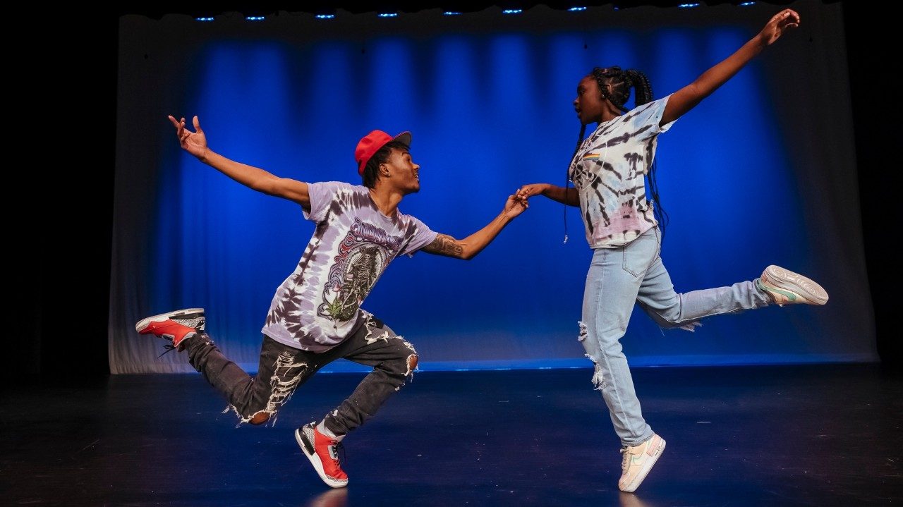  Two cast members of "Memphis Jookin': The Show" each stand on the toes of one foot. On the left is a Black man in a red hat, purple T-shirt, black ripped jeans, and red sneakers. Both of his legs are bent at right angles at the knee, almost as if he is down on one knee, but he's only on one foot. His right arm is extended out back behind him, and his left holds the hand of a Black woman with long braids, tie-dyed T-shirt, light ripped jeans, and white sneakers. She is on the toes of her left foot; her right is extended behind her, as is her left arm.