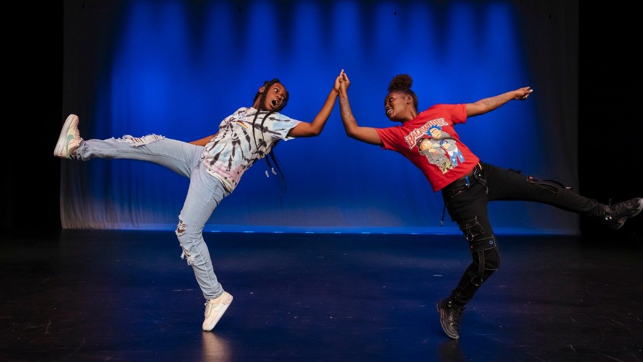  Two cast members of "Memphis Jookin': The Show" each stand on the toes of one foot. On the left is a Black woman with long braids, a tie-dyed T-shirt, light ripped jeans, and white sneakers. She holds hands with another Black woman in a red T-shirt and black jeans and sneakers with natural hair pulled into a ponytail on top of her head. They both face their bodies away from each other, but look back to make eye contact with each other, their opposite legs extended out in front of them.