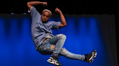  Dancer Lil Buck, a Black man with short natural bleached hair, a blue T-shirt, light ripped jeans, and black sneakers, dances on stage. He raises both arms up to flex his biceps. He is suspended in midair, his right leg extended out straight towards his left; his left leg is bent at a right angle at the knee in front of his right leg. The background is lit in blue light.