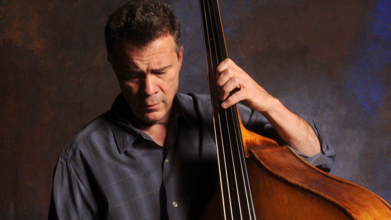  Bassist Edgar Meyer, a white man with brown hair, stands in front of a brownish blue backdrop in a grey button down shirt while playing the bass. He is looking down at the strings.