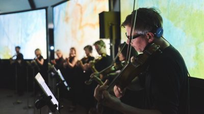  Members of the Scottish Ensemble play their instruments in a line in front of several screens, onto which art is being projected. The angle of the photo is from the side looking down the line of musicians.