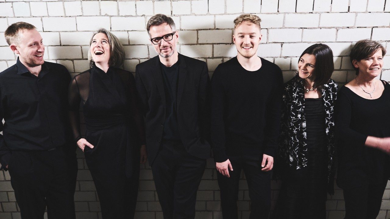  The musicians of the Scottish Ensemble stand in front of a white tile wall. The members are all dressed in black; three are white men, three are white women
