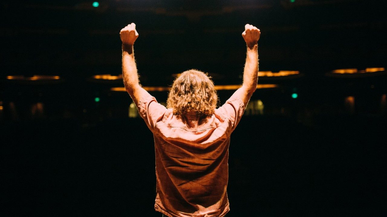 A white man with reddish brown shoulder length hair and a pink button down short sleeved shirt stands on stage facing a dark auditorium with both his fists raised in the air