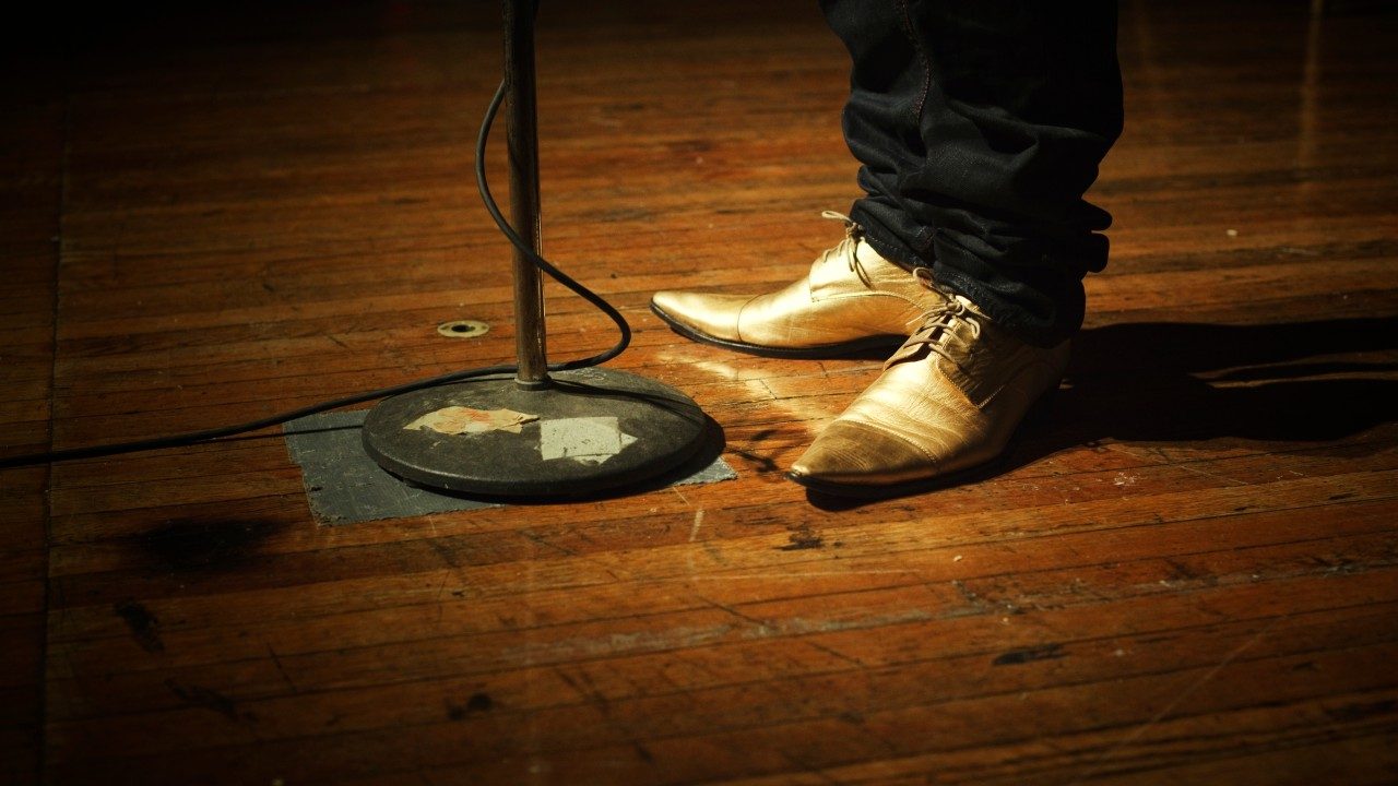  The bottom of a black microphone stand on a weathered wood floor with a person wearing pointy gold shoes and skinny black pants 