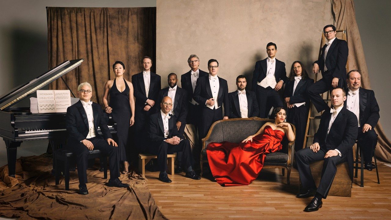  The members of Pink Martini pose in black formal wear, and China Forbes poses on a grey settee in a red ball gown.