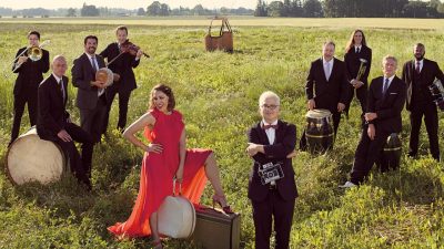  The members of Pink Martini with their instruments and vintage suitcases in an open field with the basket of a hot air balloon and a line of trees in the background. All the men wear black suits, and China Forbes wears a floor-length red dress. 