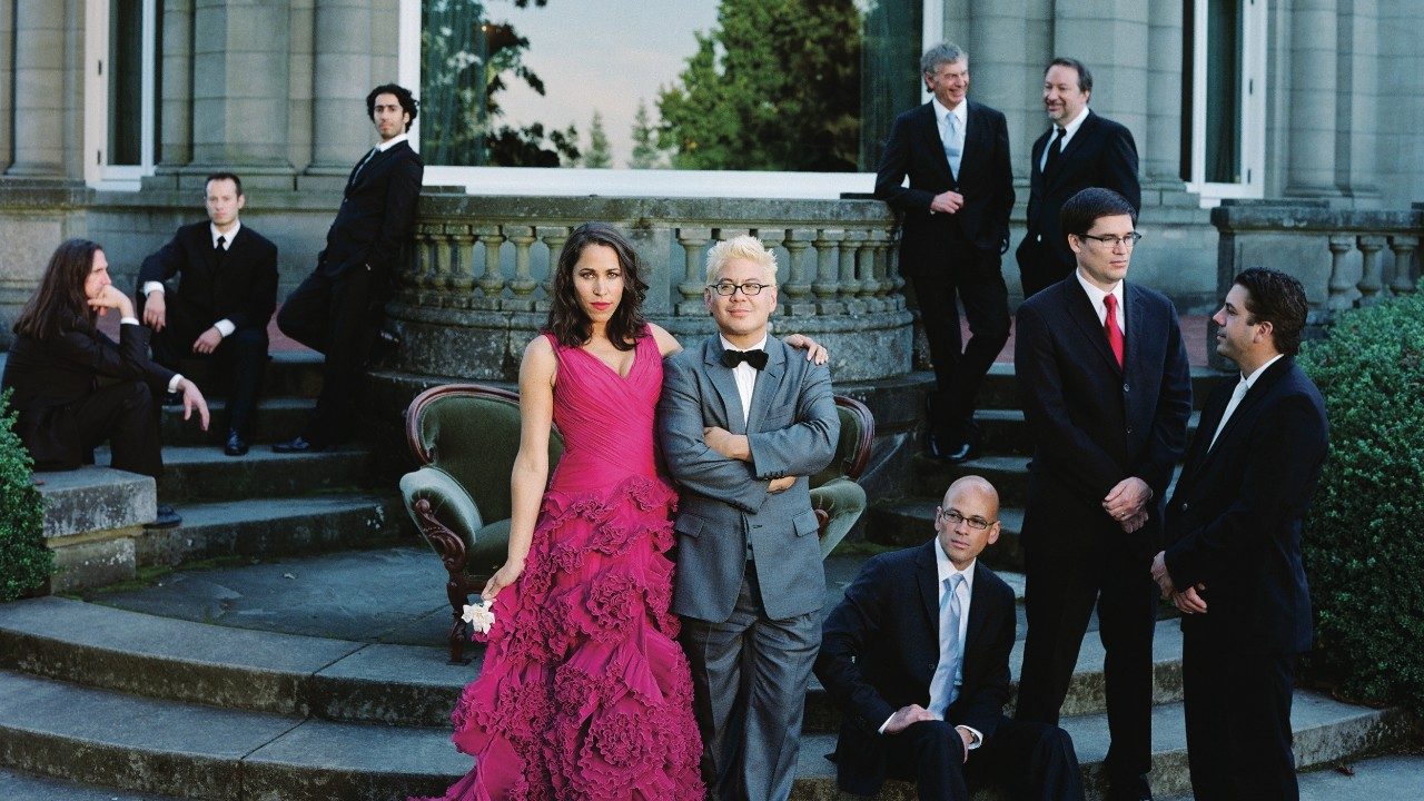  The members of Pink Martini pose on the concrete patio steps of a grand house. The men wear black suits, bandleader Thomas Lauderdale wears a grey suit, and soloist China Forbes wears a flowing, floor length hot pink ballgown.