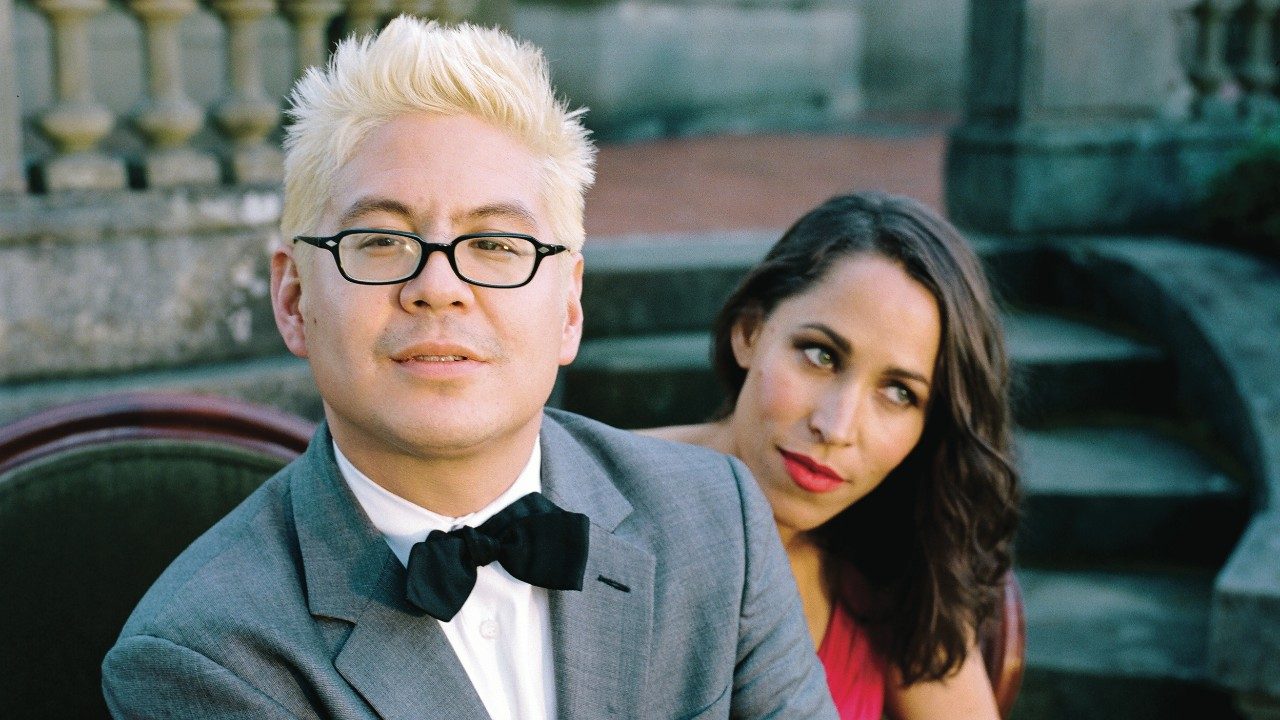  Thomas Lauderdale and China Forbes of Pink Martini on an olive green love seat in front of a large house. Lauderdale, an Asian man with bleached blonde hair and black framed glasses, wears a grey suit and black bowtie. Forbes, a white woman with green eyes and medium length dark brown hair, wears a hot pink ballgown and pink lipstick.