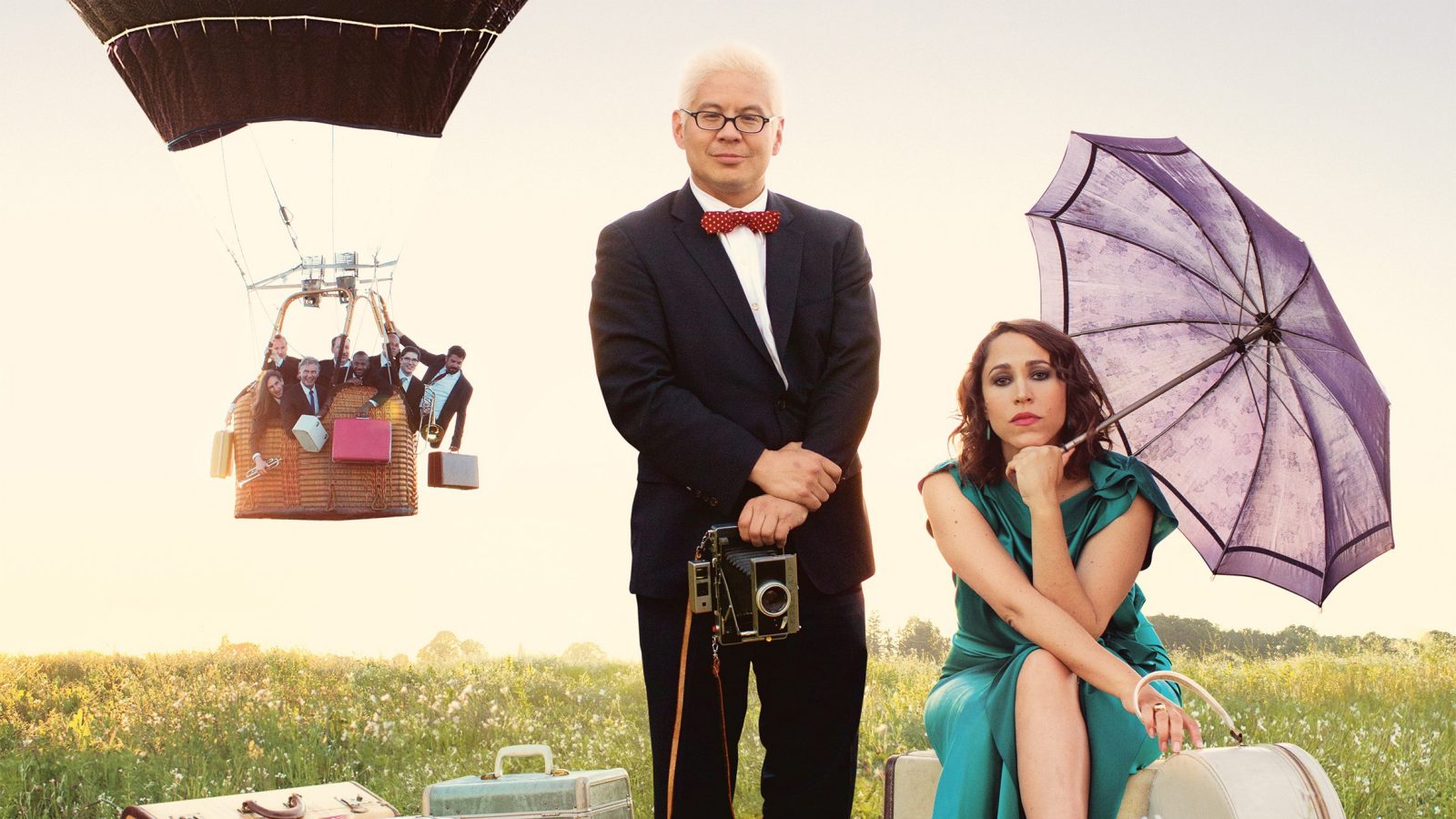  In the foreground, Thomas Lauderdale of Pink Martini, standing and holding a vintage camera, and China Forbes of Pink Martini, seated on and near vintage suitcases and holding a navy blue umbrella, while others float in a hot air balloon in the background