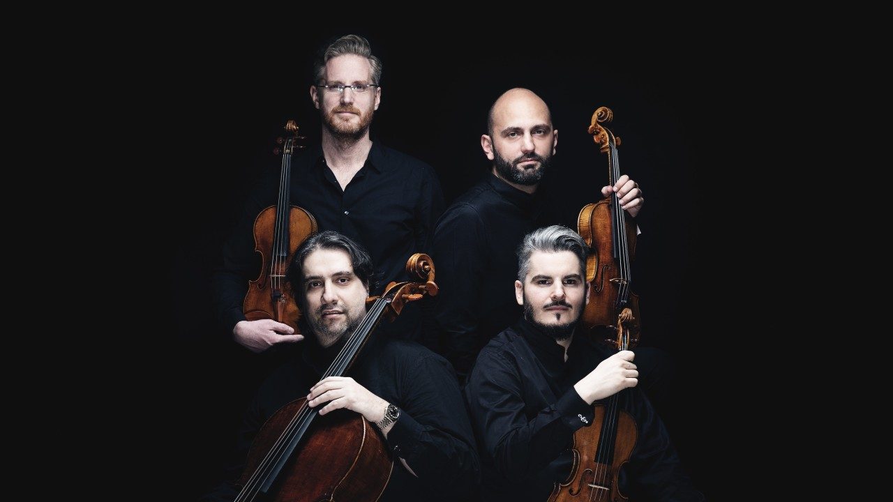  The members of Italian string quartet Quartetto di Cremona wear all black in front of a black background and hold their instruments. The quartet is made up of four white men.