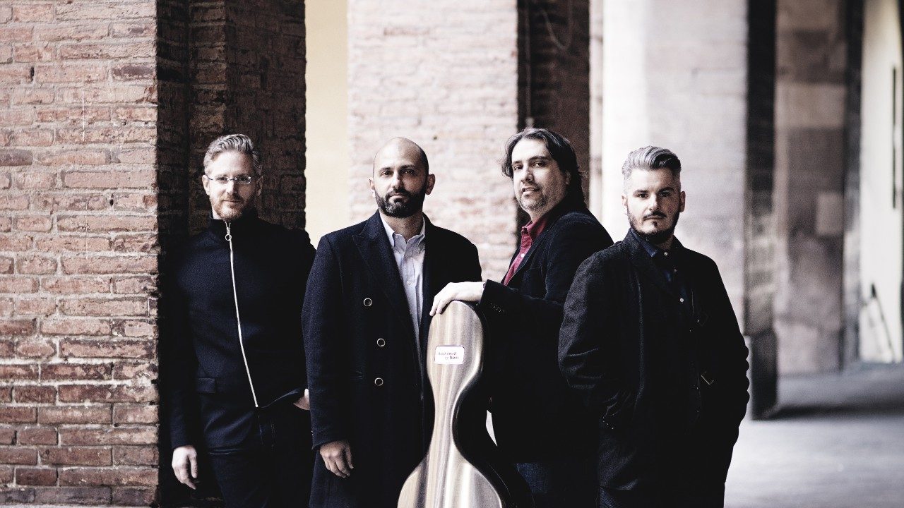  The members of Italian string quartet Quartetto di Cremona stand outside in front of brick columns. The wear black overcoats, and one holds his bass case. The quartet is made up of four white men.
