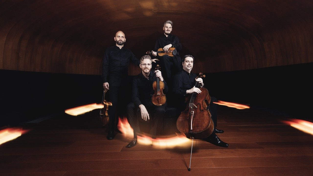  The members of Italian string quartet Quartetto di Cremona sit and stand on a stage, holding their instruments. The quartet is made up of four white men wearing all black.
