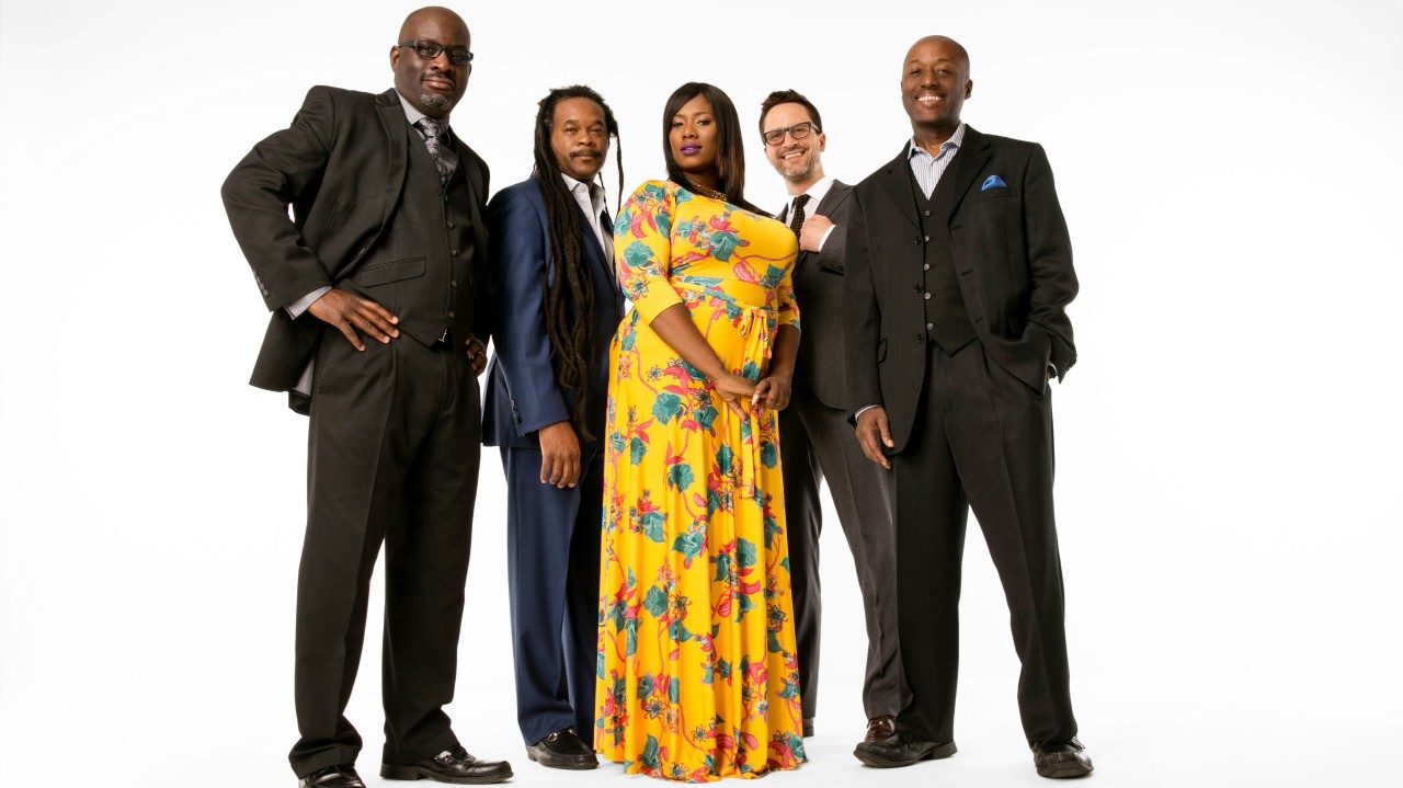  Charleston five-piece band Ranky Tanky stands in a line against a white background. The group includes four men, three of which are Black and one white, and a Black woman. She stands in the middle of the men, who wear three-piece suits in shades of blue, grey, and black, wearing a floor length yellow floral dress.