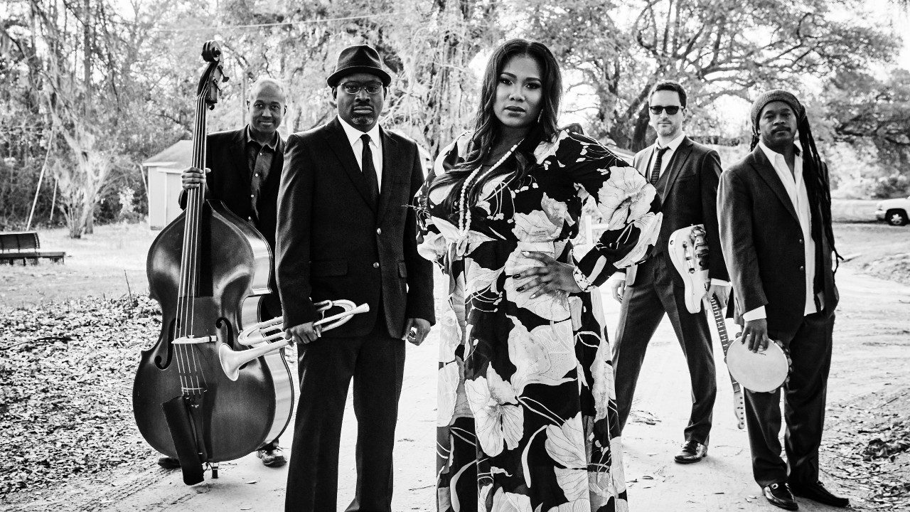  The members of Ranky Tanky stand on a sandy dirt road, presumably in or near Charleston, South Carolina, in this black and white image. The lead singer, a Black woman, stands in the center with one hand on her hip, wearing a floor length long-sleeved floral dress. The band members, three Black men and one white man, hold their various instruments: bass, trumpet, electric guitar, and tambourine.