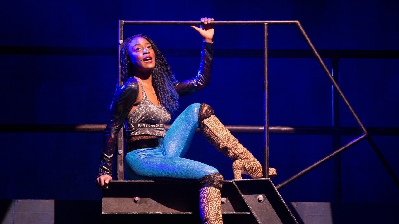  A cast member of "Rent" sings on stage. She is a Black woman with long braids parted in the middle. She is wearing a grey tank top, black shrug, tight blue pants, and leopard print knee-high boots. She sits on a prop fire escape with one knee bent up towards her chest and one hand holding a metal railing.
