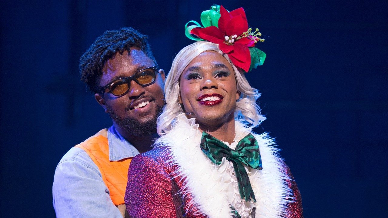  Two cast members of "Rent," both Black people, smile towards the camera. The one in front wears a Mrs. Claus suit, blonde wig, and poinsettia hat, and the other embraces them from behind and wears a blue button down shirt, orange vest, and brown glasses.