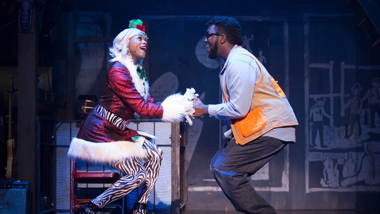  Two cast members of "Rent" sing and dance on stage, both bent at the knees and holding hands facing each other. They are Black people, one wearing a Mrs. Claus outfit, zebra print leggings, and blonde wig, the other wearing a blue button down shirt, orange vest, and grey pants.