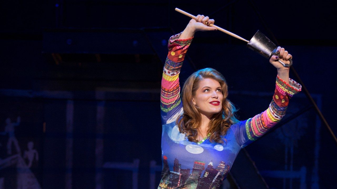  A cast member of "Rent" holds a cowbell above her head and hits it with a drumstick. She is a white woman with light brown hair, red lipstick, and a multicolored patterned shirt.
