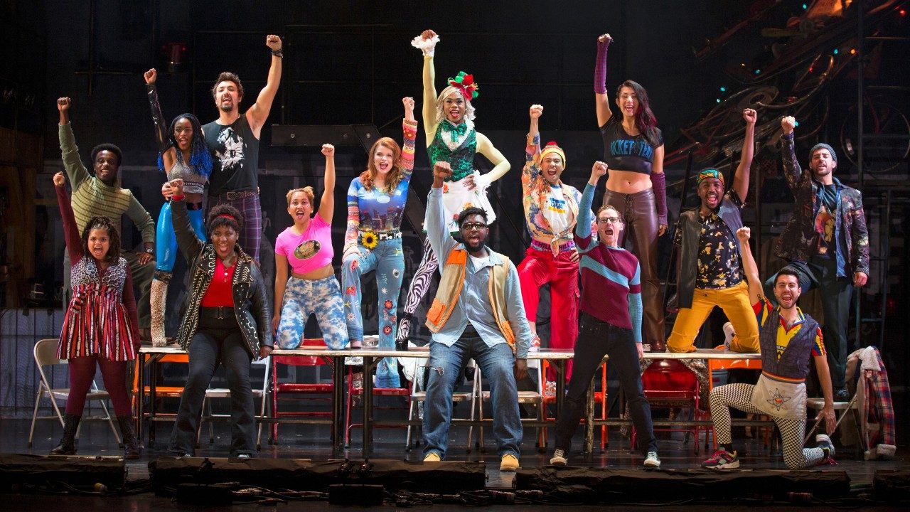  The cast members of "Rent" sing and dance on stage, all facing the audience and all with one fist raised above their heads