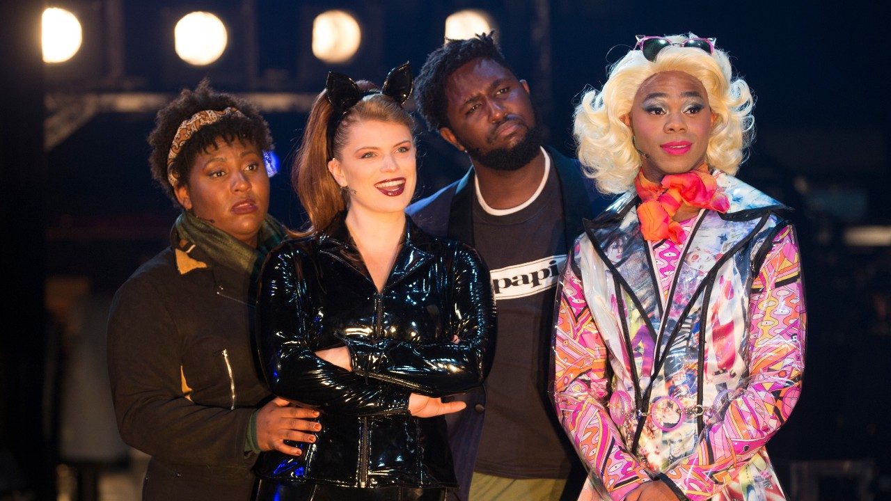  Four cast members of "Rent" stand in a row. They are judging something. From left to right is a Black woman in a green scarf and black jacket, a white woman in a black leather catsuit with cat ears, her light brown hair pulled into a ponytail, a Black man with a black jacket and a black T-shirt underneath that reads "papi," and a Black person with bleach blonde wig, pink and orange scarf, and multicolor jacket.