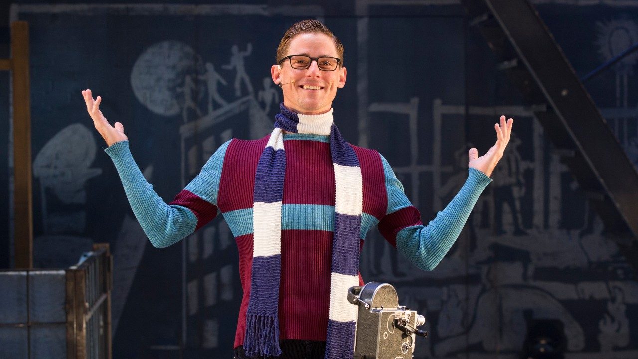  An actor from "Rent" stands on stage, smiling towards the camera, with his hands thrown up in a shrug. He is a white man with brown hair and black rimmed glasses,  wearing a sweater and a scarf.