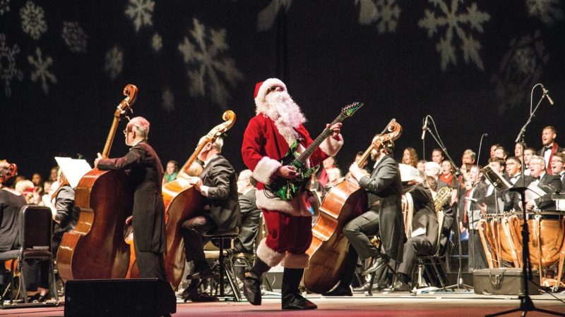  The Roanoke Symphony Orchestra performs "Holiday Pops Spectacular" at the Moss Arts Center. In the center of the image is a guitarist dressed in a Santa costume playing a green electric guitar. The rest of the orchestra surrounds him, and lights on the back wall look like snowflakes.
