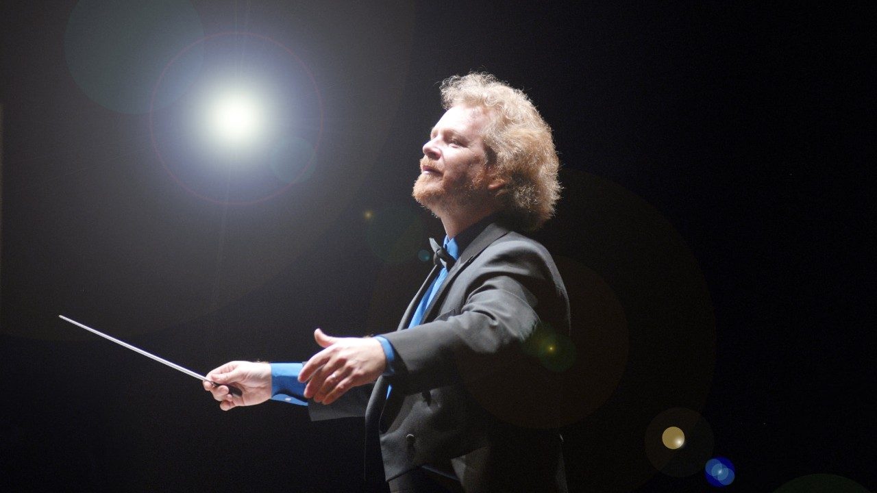 Conductor David Stewart Wiley, a white man with wavy blonde hair and a blonde trim beard, conducts in a black suit jacket and cerulean blue shirt and bow tie. Behind him is a black background except for one bright light shining on him.