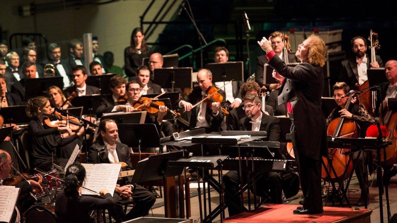  David Stewart Wiley, a white man with wavy blonde hair, a black suit, and a red shirt, conducts the Roanoke Symphony Orchestra from a red riser.