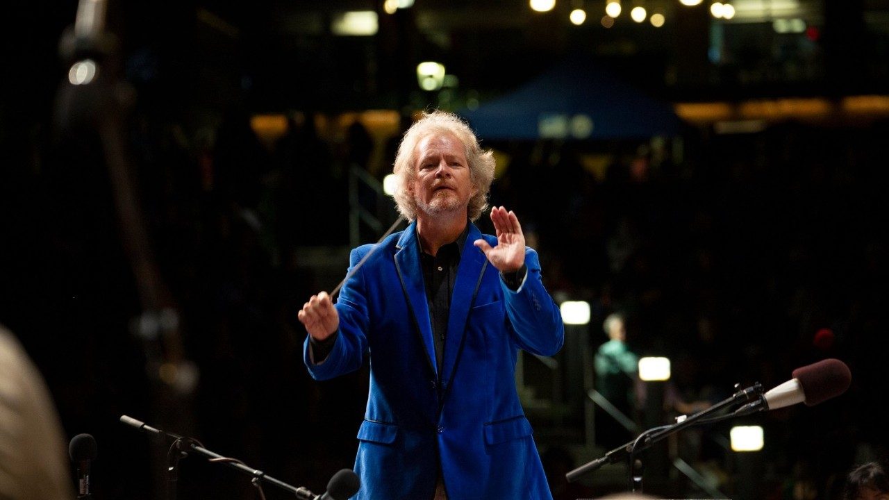  David Stewart Wiley, a white man with wavy blonde hair and a velvet blue jacket and black shirt, conducts the Roanoke Symphony Orchestra. Behind him is the empty part of a stadium and several lights. He is facing the camera; the photo was taken from behind the orchestra.