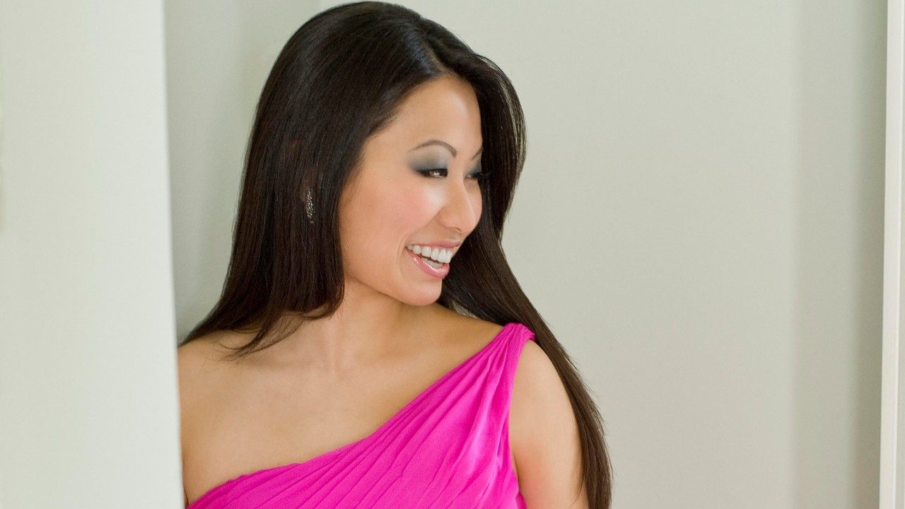  Violinist Sarah Chang, an Asian woman with long dark brown hair, smoky eye makeup, and nude lipstick, wears a hot pink one-shoulder dress and leans against a beige wall. She is smiling at something to the right of the camera.