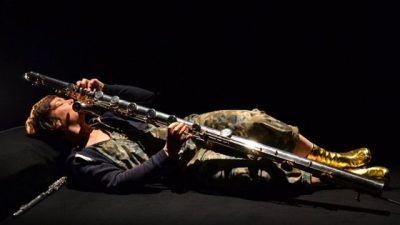  Flautist Claire Chase reclines on a black chaise lounge against a black background while playing a flute. She is a white woman with short brown hair wearing a dark blazer over a green and brown patterned jumpsuit and shiny gold boots.