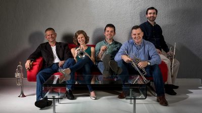  The members of wind quintet Fifth Bridge hold their instruments and sit on a red couch in front of a textured grey wall and behind a glass and metal coffee table. The members are four white men and one white woman.