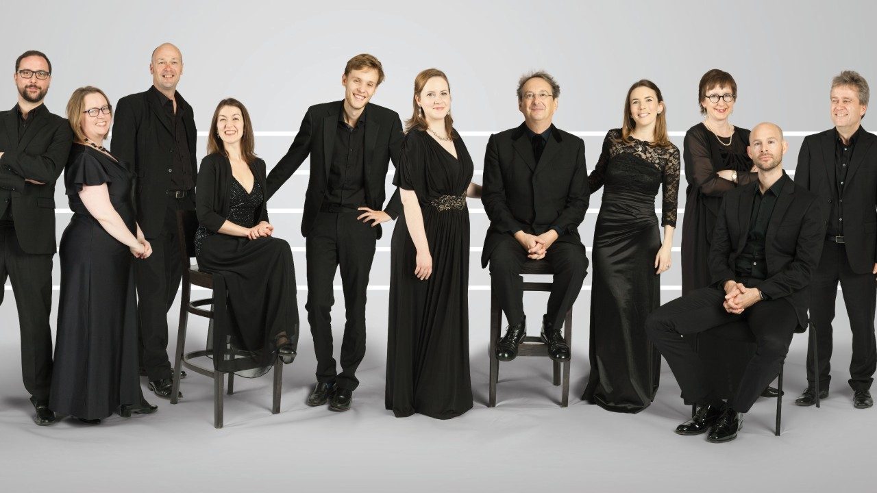  Members of the Tallis Scholars stand and sit on stools in front of a grey backdrop. The group is six white men and five white women. All members are wearing all black.