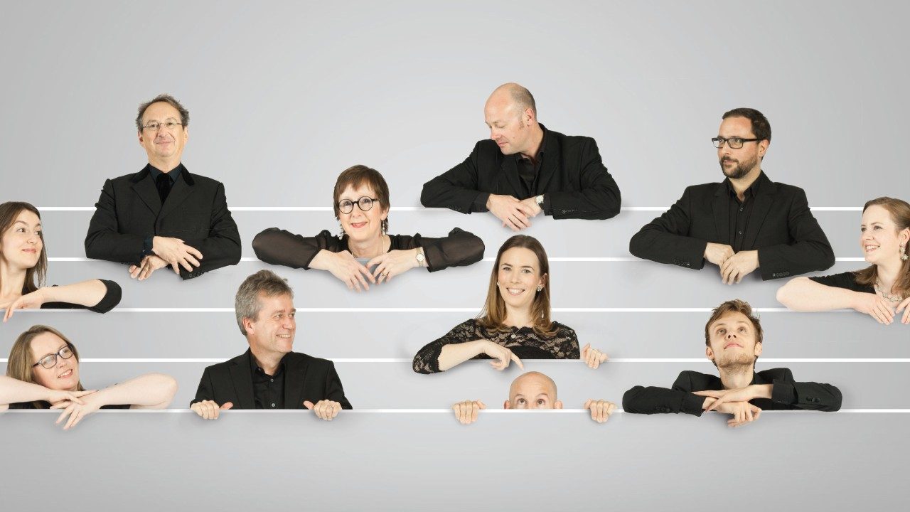  The members of the Tallis Scholars appear to peek above lines on a music sheet. All are wearing black; there are six white men and five white women.