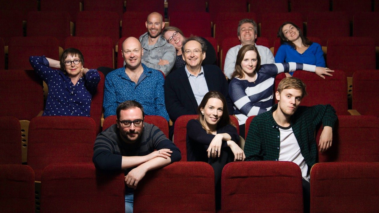  Members of the Tallis Scholars sit in red auditorium seats. The group is composed of six white men and five white women. They're wearing day wear. Three members are in the first row, four in the second row, and four in the back row.