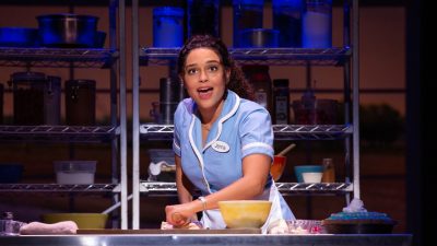  Jisel Soleil Ayon as Jenna. Jisel is a Black woman with natural hair wearing a powder blue diner uniform. She rolls out a pie crust while she sings. Behind her are metal industrial kitchen shelves filled with pantry items, and on the counter in front of her is a yellow blow and a pie with blue icing and whipped cream.