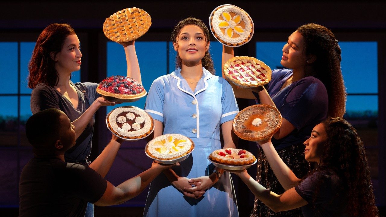  Jisel Soleil Ayon, a Black woman with dark curly hair worn half up and wearing a blue diner uniform, stands in the middle of four people holding eight pies around her in an oval shape.