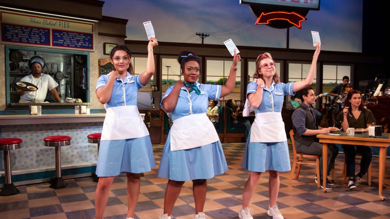  Three women actors in "Waitress" stand in a diner wearing powder blue diner uniforms and holding meal tickets in their left hands above their heads.