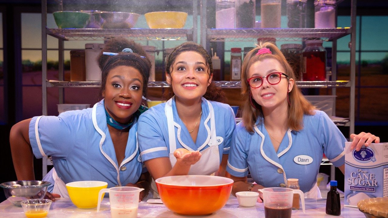  Three women actors in "Waitress" smile towards the camera wearing powder blue diner uniforms and white aprons. In front of them is a counter filled with the items needed to make a pie, and behind them is a metal kitchen storage shelf filled with pantry items. The middle woman tosses a bit of flour up into the air.